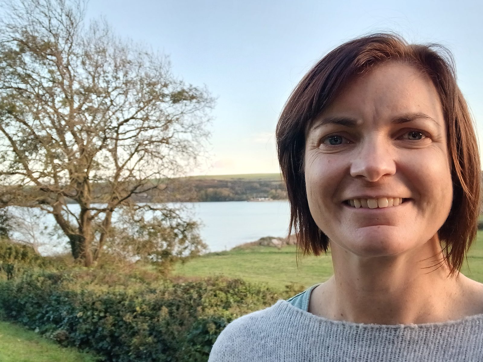Cecily Maher smiling outdoors, with a tree, green fields and water behind her